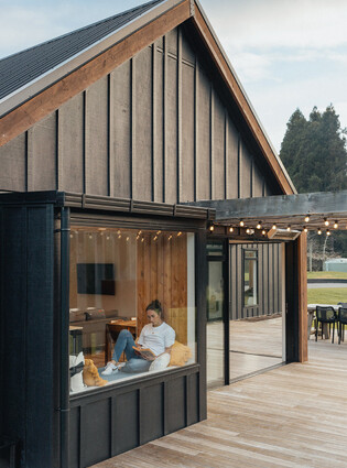 This image shows the completed home with a deck, outdoor fairy lights on the pergola, a modern board-and-batten home, and the homeowner reading a book in the window seat.