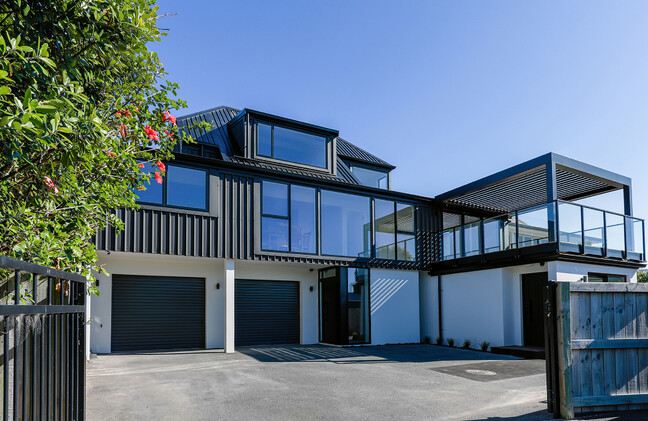 The image shows a newly completed builder's renovation in Papamoa. The home is two two-story with white textured coating and a twin garage on the lower level, and on the upper level, a new deck with pergola, profiled black metal cladding, and large windows. The image is taken from the driveway.