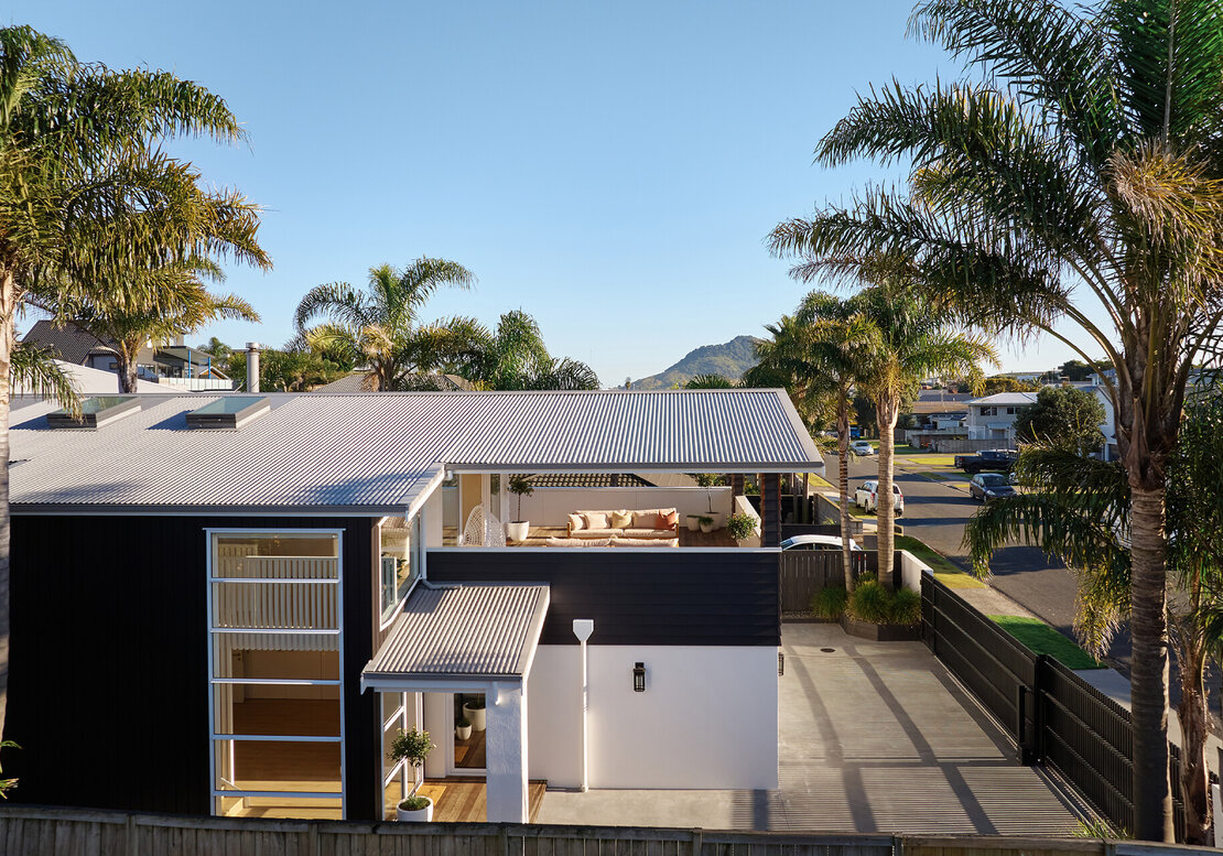 A full home renovation in Mount Maunganui, showing a modern home with a covered deck, surrounded by palm trees, with Mount Maunganui (Mauao) in the distance.