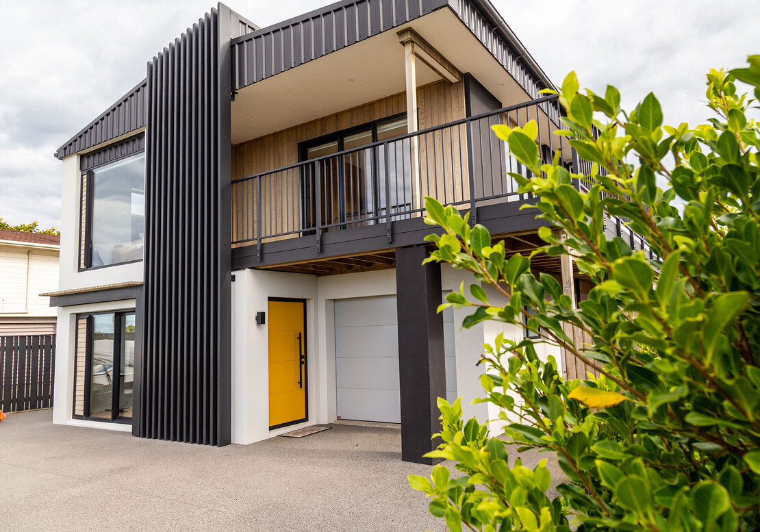 The image shows a recent renovation completed in Papamoa. It's a modern two-story house with a yellow front door, new cedar cladding and cream textured coating, and a deck on the second story.