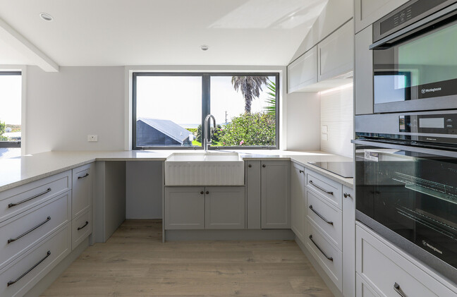 A newly completed kitchen renovation in Papamoa. A grey country-styled kitchen and wood-look laminate flooring. The kitchen has a new ceiling and new joinery. The large kitchen window looks out onto the ocean view at Papamoa Beach.