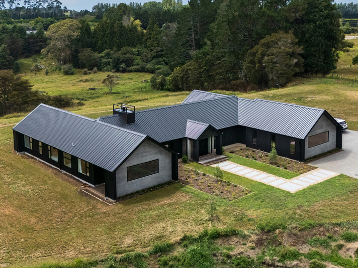 The project cover image shows a large new home built in rural Katikati. This new build shows a high level of workmanship completed by Straight Up Builders Tauranga. The image shows the exterior of the home, with wood-grain-effect concrete panels and black cladding, a new front door, and a Colorsteel roof.