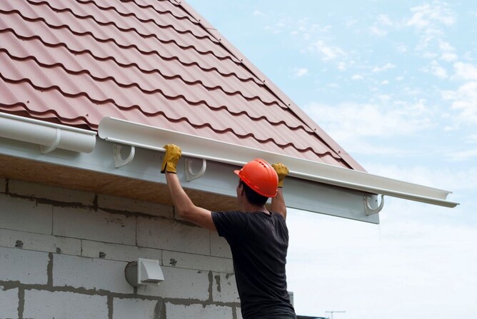 Roofer installing new white spouting on a residential home with red metal roofing.