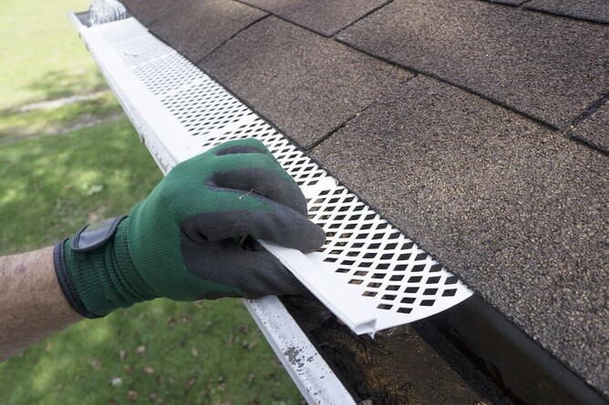 A man with green work gloves installing a white plastic mesh gutter guard on a shingle roof to prevent leaf and debris build-up.