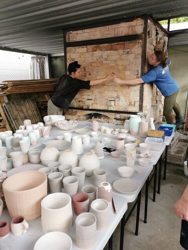 Alex and Larry stand on 2 sides of the bricked kiln. Alex passes tape to Larry