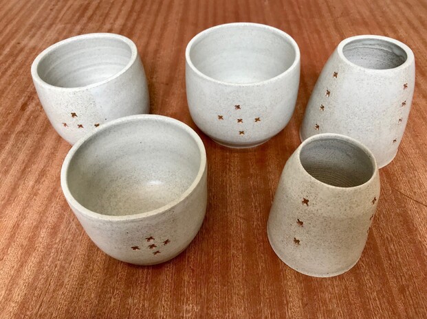 A collection grey ceramic ware sits on a brown wooden surface. The 5 ceramic vessels are decorated with brown crosses