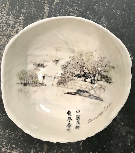 White ceramic bowl that is handpainted with black underglaze . Painting shows a scene with trees, boats, and a wall and has some Chinese characters. It is signed "Sandra Lin"