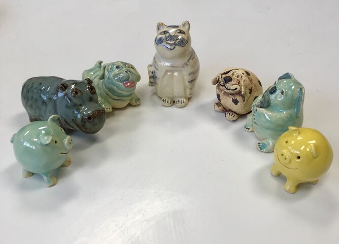 a group of 57 ceramic animals in a semicircle: left to right: a turquoise pig, a blue hippo, a turquoise pug dog, a white cat, a brown dog, a turquoise koala bear and a yellow pig