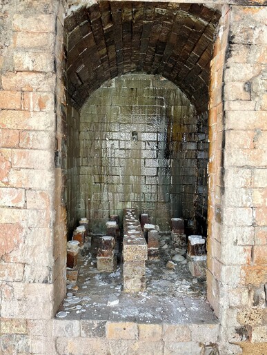 An empty kiln with some stacks of bricks and broken ceramic shards sitting on the bottom. The back of the kiln has a wet appearance with a forest green hue.