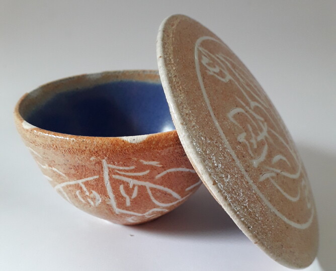 Ceramic bowl with lid. The lid is open and leaning to and against the right side of the bowl. The bowl is blue on the inside and orange on the outside with etched white sgraffito patterning