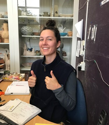A smiling young wahine sits at a desk gesturing thumbs up with both hands