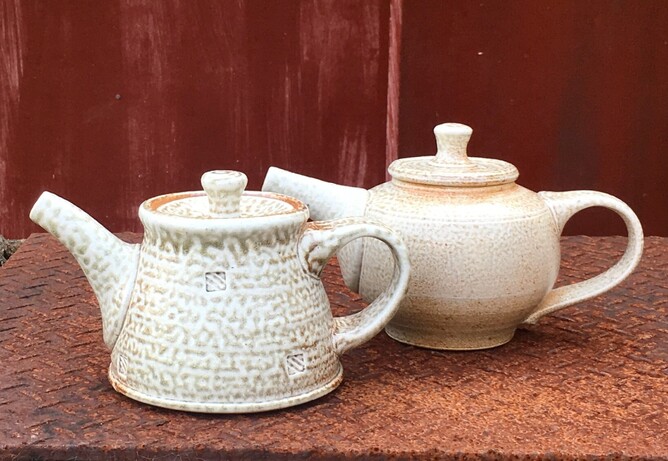 Two off white ceramic teapots sit on top of a rusted metal surface