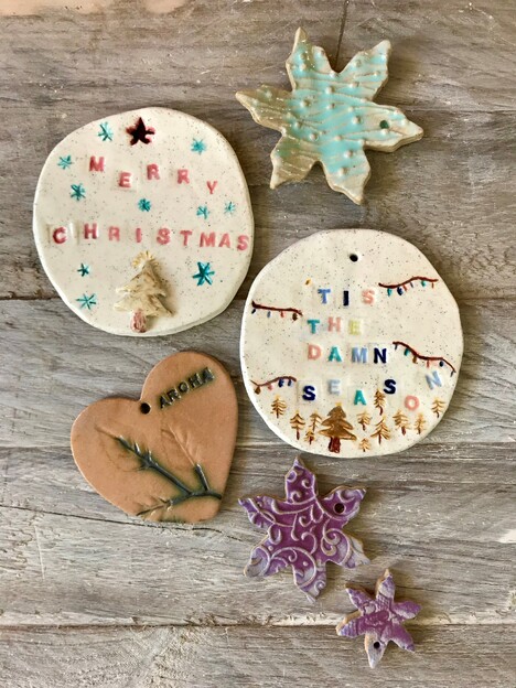 6 ceramic Christmas decorations on top of a rustic wooden tabletop. A turquoise snowflake, two circular decorations: one with the words "Merry Christmas" and another reads "Tis the damn season", a heart and two purple snowflakes