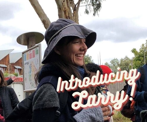A woman wearing a dark blue hat stands below a tree, she holds a cup in her hand and has a broad smile on her face. Text on the image reads "Introducing Larry"