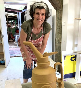 A woman with a pale coloured headband is smiling at the camera, bending forwards over a ceramic teapot in the foreground. She wears an olive green tee shirt and jeans with a red and white striped apron on top.