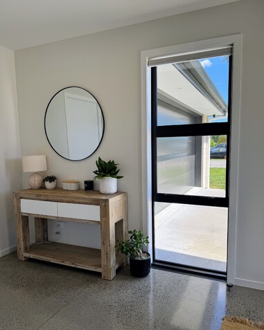 Image of front entry of new home. Sunlit with tiled flooring. Styled with light timber credenza and circle mirror. Home built by Coull Builders, Otorohanga.