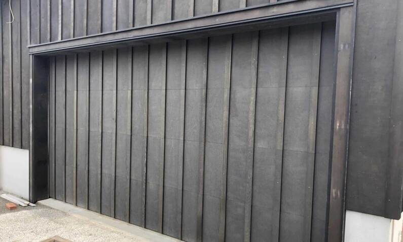 Photo of a black plywood/board and batten garage door. Panels are arranged in a vertical pattern, repeating across the length of the garage matching the exterior walls of the house.