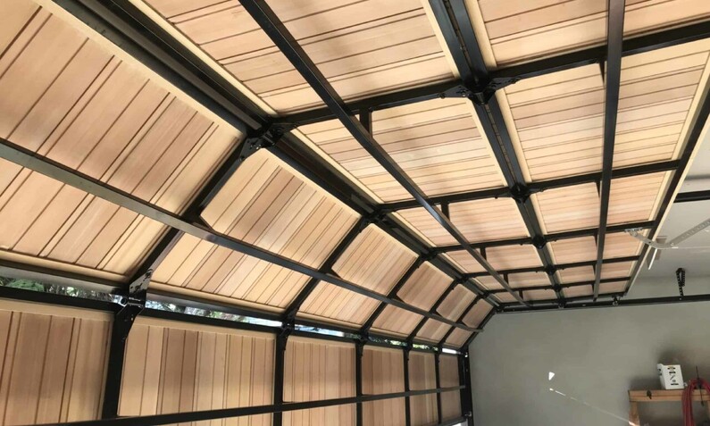 Photo taken from inside a garage, showing a timber, wooden sectional garage door opening. Timber is sand coloured and arranged in a vertical pattern. Horizontal steel beams divide the garage into four sections