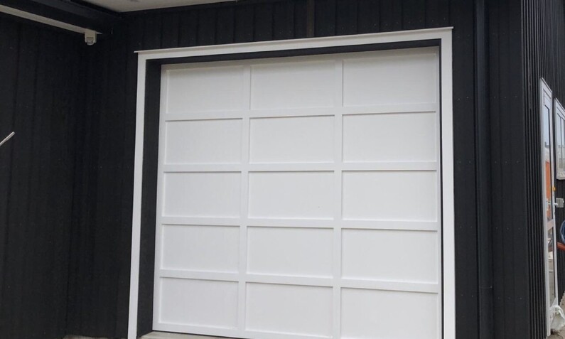 Photo of a white plywood/board and batten panel single garage door fitted to a Rodney, Auckland home. Boards are in a 3 x 5 pattern. White garage door contrasts the black house and matches the white trim of the house