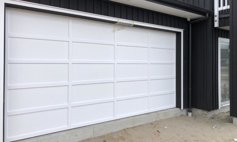 Photo of a newly installed, white plywood/board and batten panel double garage door fitted to a Rodney, Auckland home. Boards are in a 4 x 5 pattern. White garage door contrasts the black house and matches the white trim of the house.