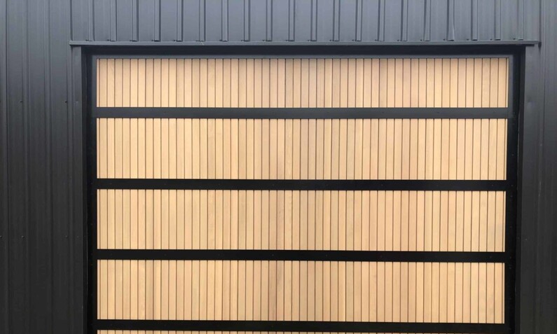Photo of a timber garage door fitted to a home in the Rodney, Auckland area. Thin wooden timber with a vertical pattern, split into four sections by black horizontal steel