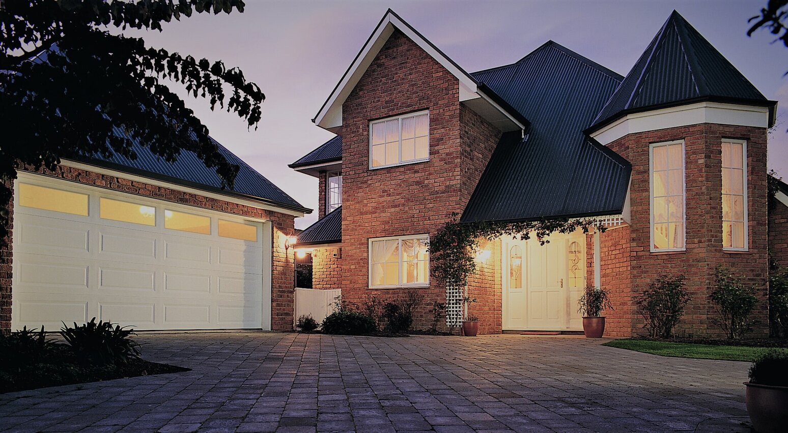 Photo of a house at dusk, lights on inside the house and visible through the garage door. Garage door is a Dominator Somerset garage, with four rectangular windows along the top panel of the door.
