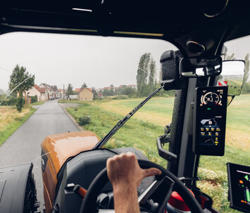 View from inside the Valtra S Series cab, showing a farmer’s hands on the steering wheel, driving down a rural road with the SmartTouch interface and expansive front visibility.