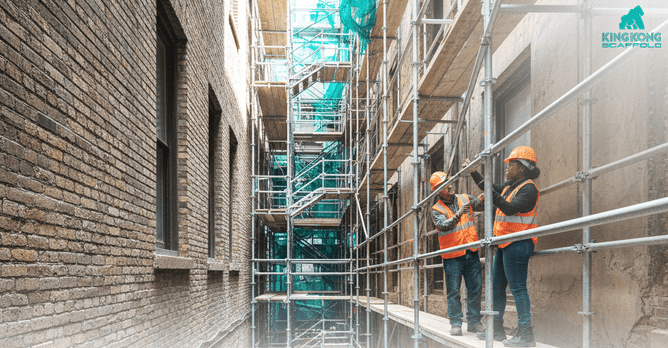 Two construction workers in high-visibility vests and hard hats installing modular scaffolding within a narrow brick alleyway between old buildings, with safety netting visible overhead.