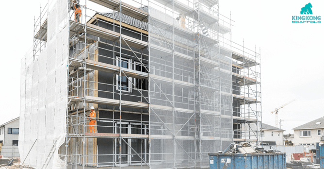 A three-story residential house under construction or renovation, fully covered in safety netting and scaffolding, with construction workers in orange vests performing exterior work.
