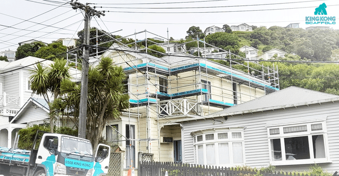 A large two-story house situated on a steep hill, fully wrapped in a secure scaffolding structure to allow for exterior maintenance or painting.
