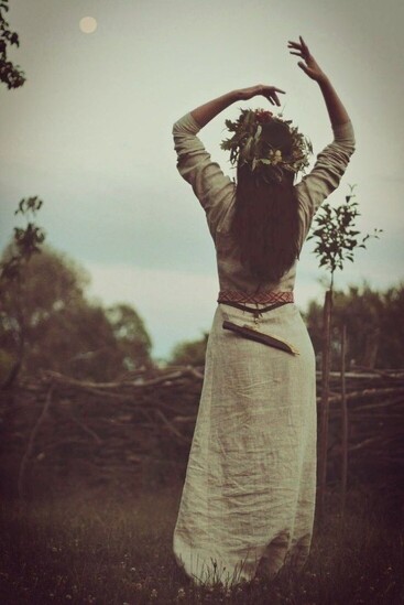 “Woman wearing a natural linen dress and flower crown, raising her arms toward the sky in a nature setting, symbolising feminine archetypal energy.”