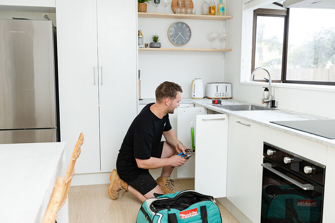 Plumber kneeling beside a white kitchen sink bench