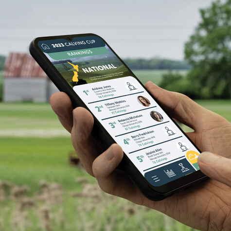 Hand holding mobile phone while using the Elanco Calving Cup app against a rural background.