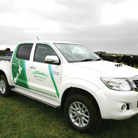 White ute with green and blue decals, map of New Zealand and Open Country logo on side door, in a paddock with cows behind.