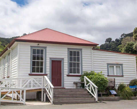 The Kūaotunu Hall as seen from the front - a white painted wooden building with a red roof and red door, with steps leading up to it and a ramp to the left