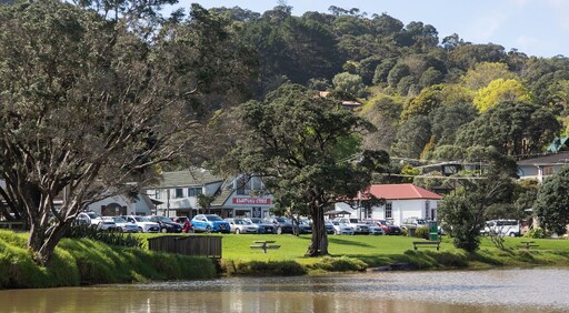 A view of the Kūaotunu Hall as seen from the other side of the Kūaotunu river. The white painted wooden hall with its red roof can be seen in the distance next to the Kūaotunu store and a row of cars parked by the riverside.
