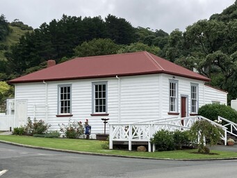 The Kūaotunu Hall in 2023 - a white wooden building with a red roof, with steps up to the front door and a ramp to the left side