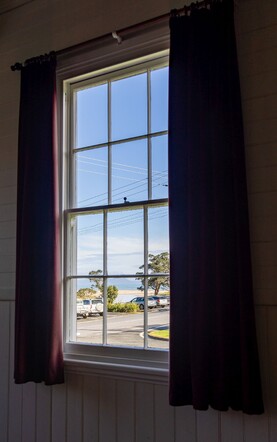 View of the Kūaotunu river and beach through one of the tall sash windows, flanked by red velvet curtains, in the historic Kūaotunu Hall