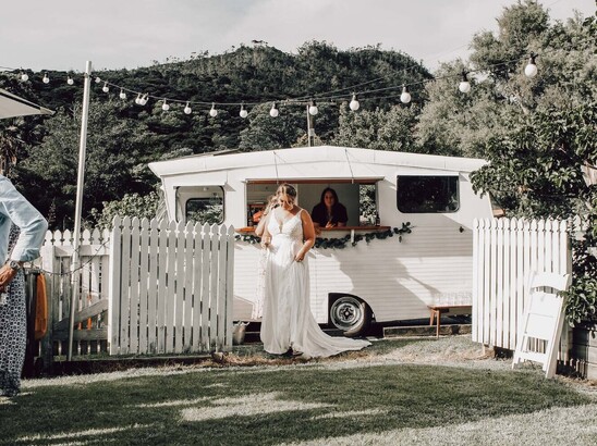 A bride with a glass of champagne standing outside a caravan bar parked by the white picket fence outside the Kūaotunu Hall