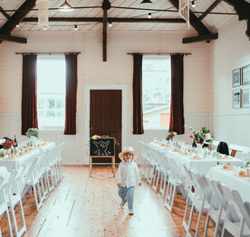 A child wearing a white hat walking between a row of wedding tables and chairs at the Kūaotunu Hall. In the background are two high sash windows with velvet curtains with a red door in between.