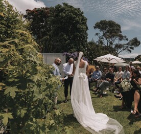 A bride and groom standing on the back lawn of the Kūaotunu Hall surrounded by wedding guests watching the ceremony
