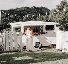 A bride with a glass of champagne standing outside a caravan bar parked by the white picket fence outside the Kūaotunu Hall