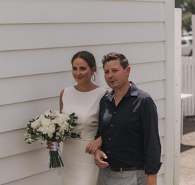 A bride carrying a bouquet of flowers walking along the white wooden south wall of the Kūaotunu Hall, escorted by her father