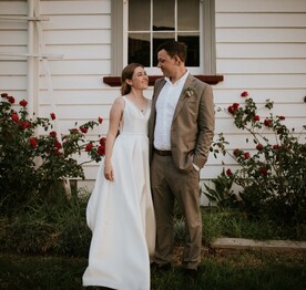 A bride standing with her groom outside the Kūaotunu Hall with a display of Dublin Bay red roses in the garden behind them