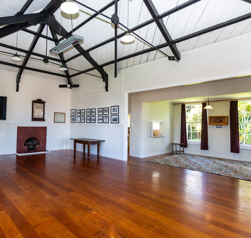 An interior view of the Kūaotunu Hall showing the main part of the hall and the annex on the right hand side. The floor of the hall is swamp kauri and the walls are white, there are two tall sash windows with red velvet curtains.