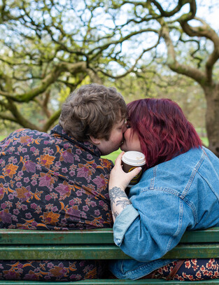 Couple share a kiss, coffee in hand during their walk through a beautiful park