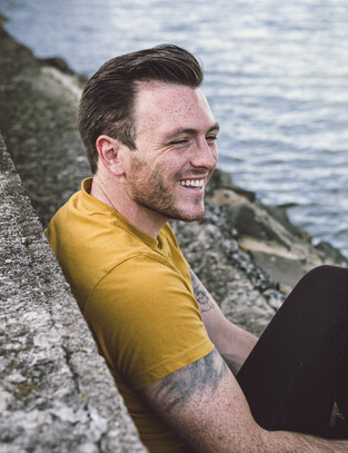 Man poses for photo, smiling and laughing with harbour behind him