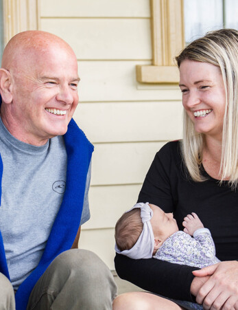 Father and daughter sit side by side smiling as the daughter holds her own newborn daughter in her arms